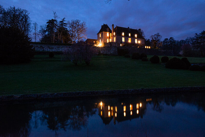 Photographie de nuit du château de Garraube toutes lumières allumées, se reflétant dans le bassin du domaine